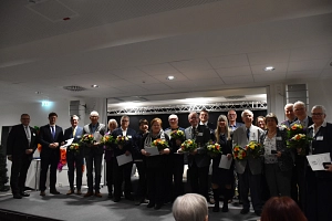 Träger:innen der Ehrennadel - Verleihung 2026 Die Träger:innen der Ehrennadel stehen mit ihren Urkunden und Blumensträußen in der Hand auf der Bühne.